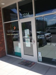The entrance to Spark Center has a single door with its handle on the right side. On either side of the door are large and full windows. On the left window is the logo of the Spark Center. Above the door is etched the address number.
