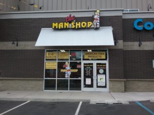 The entrance to the Man Shop has large glass windows to the left of the double doors. The doors will open outward when pulled. There is an awning above the doors and front window. The business hours are printed on the left side door. The logo is printed on the window to the left of the doors.