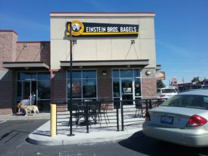 The main entrance to Einstein Bros is on the left. The exit door has printed upon it an arrow pointing to the left and to the entrance. Both entrance and exit are underneath awnings.